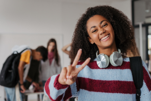 A happy, smiling teenager wearing headphones and a backpack makes a peace sign, with other students in the background