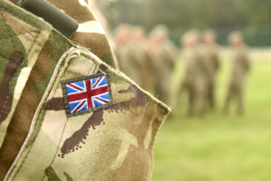 Close up of a camouflage military uniform sleeve with a Union Jack patch, with blurred soldiers in the background