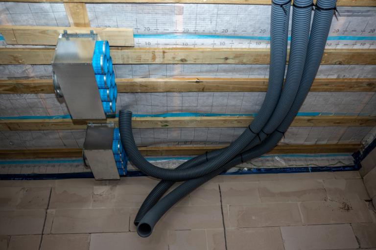 Heat recovery ventilation system with metallic boxes, blue connectors, and black ducts on wooden framework in a partially constructed home.